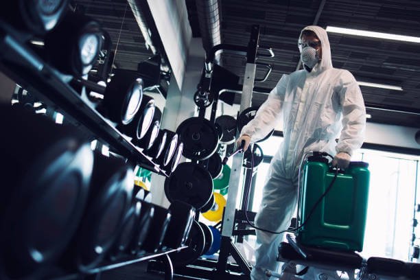 Gym disinfection and healthcare. Man in white protection suit disinfecting and fitness equipment and weights to stop spreading highly contagious coronavirus or COVID-19.